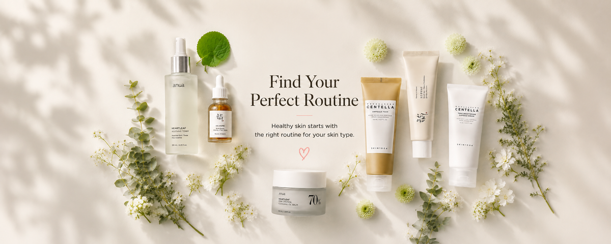 A flat-lay arrangement of multiple skincare products on a bright white background with soft leaf shadows. The products include bottles, tubes, and jars in white, beige, and amber tones, styled with small green flowers and sprigs.